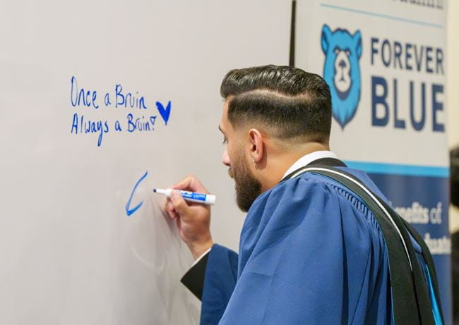 A student writes a message in blue marker on a whiteboard. The board reads, Once a Bruin, always a Bruin.