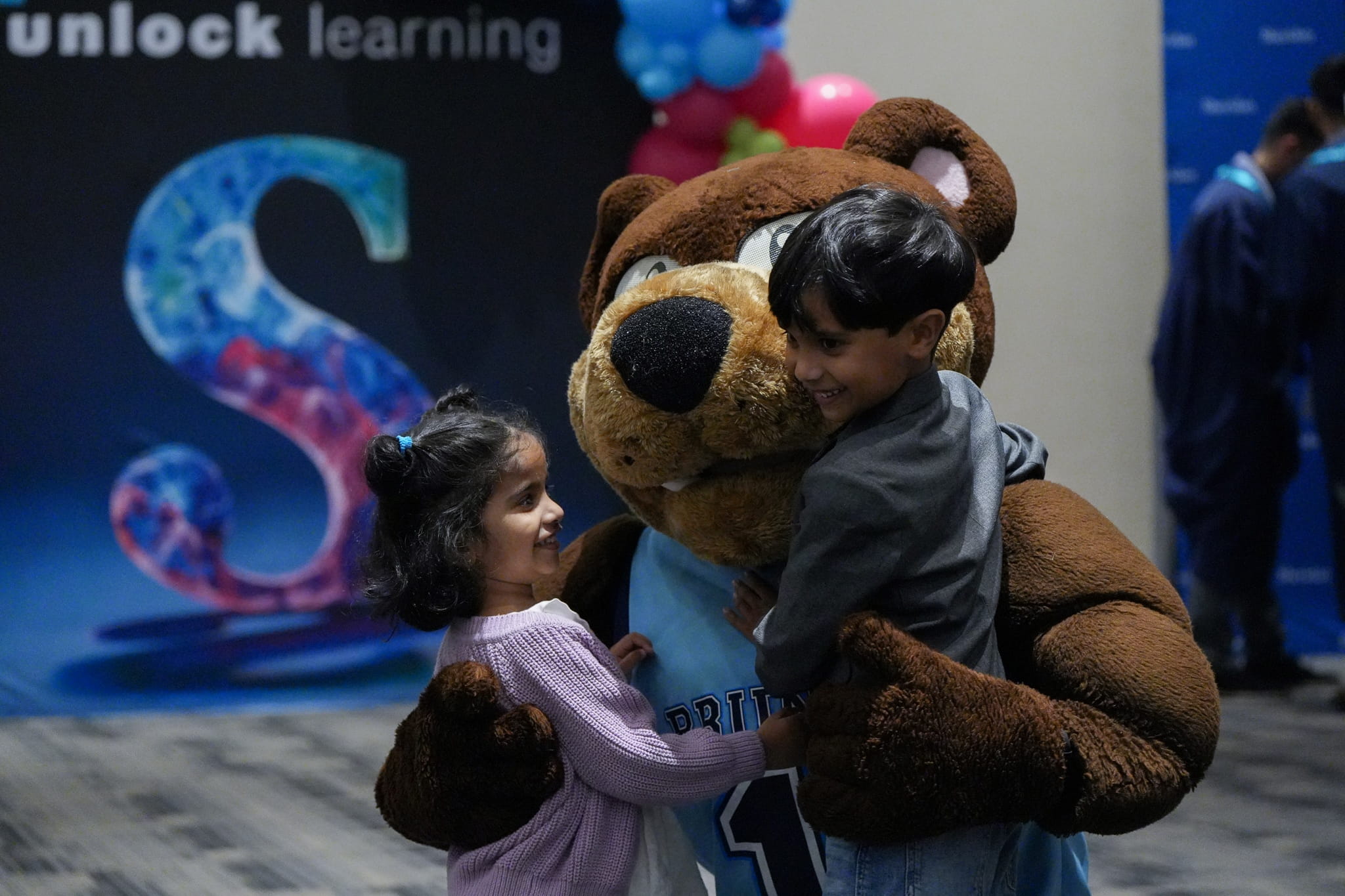 Mascot Bruno the bear crouches down to hug two children, who are smiling.