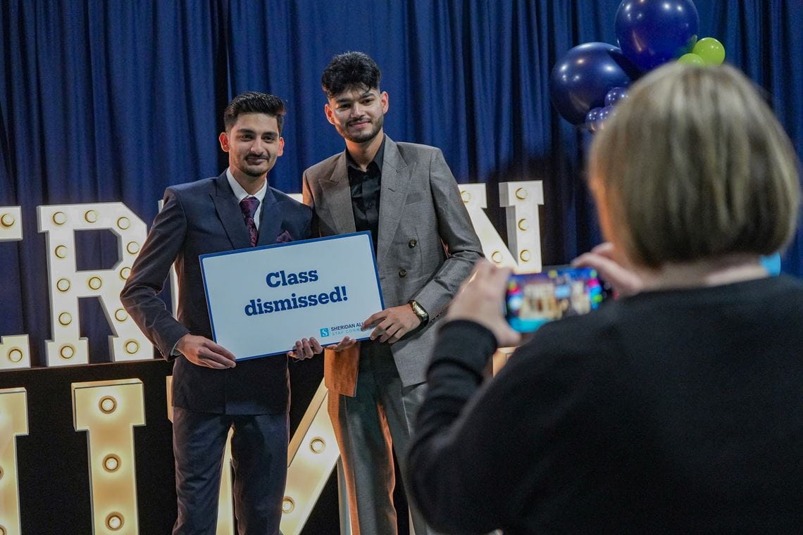 Two male students hold a sign that says class dismissed, while they have their photo taken.