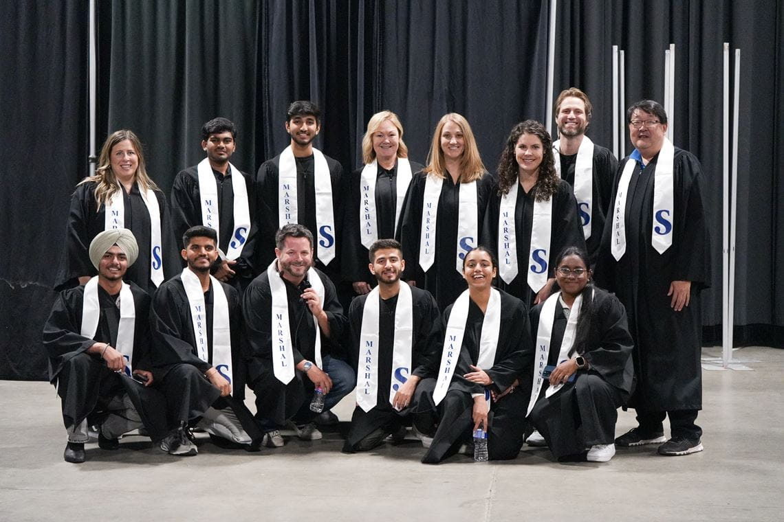 Fourteen Sheridan staff and students wear black robes and a sash that says marshal.