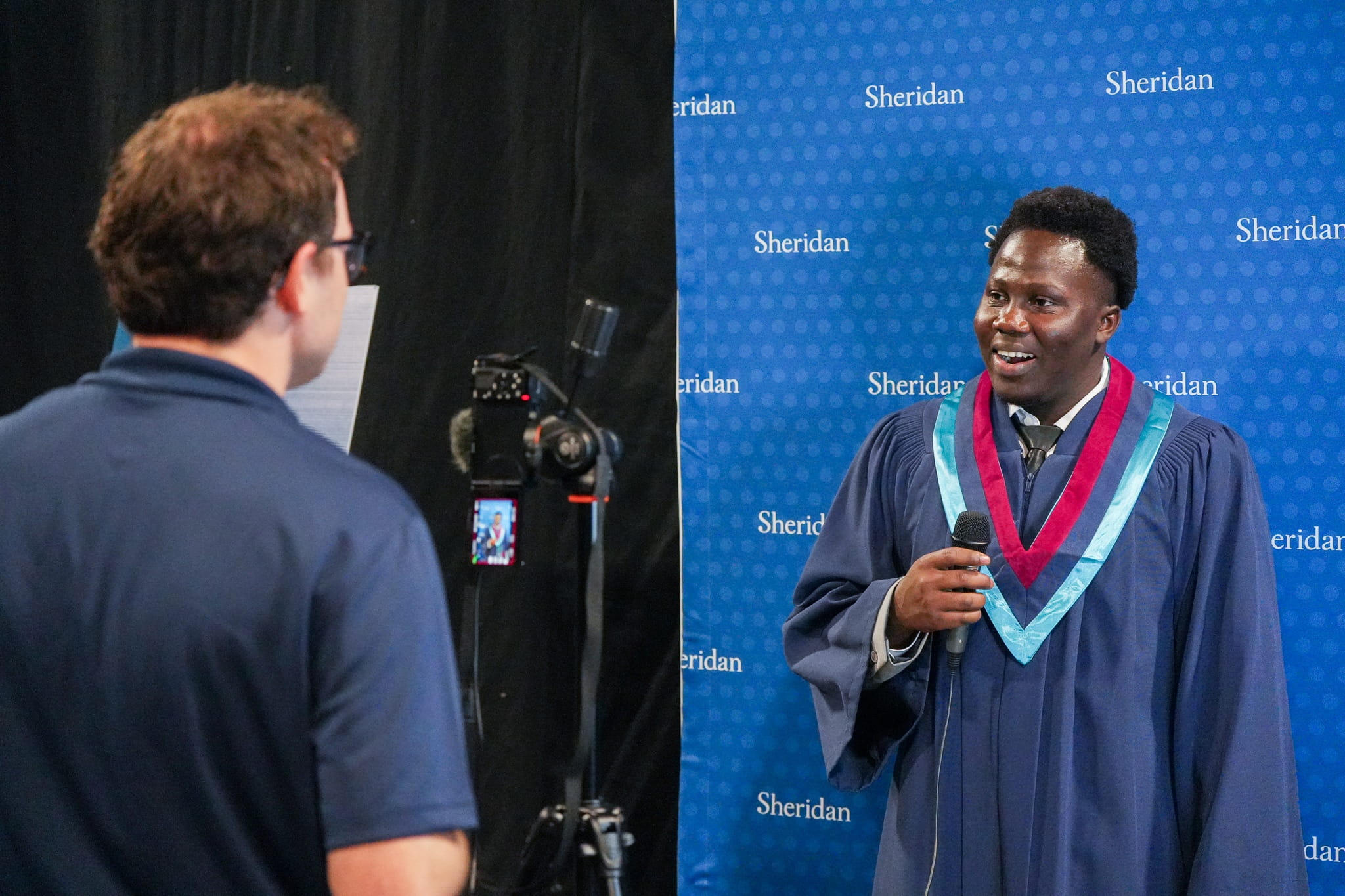 A Sheridan staff member speaks to a Sheridan graduate who is speaking into a microphone in front of a blue Sheridan backdrop.
