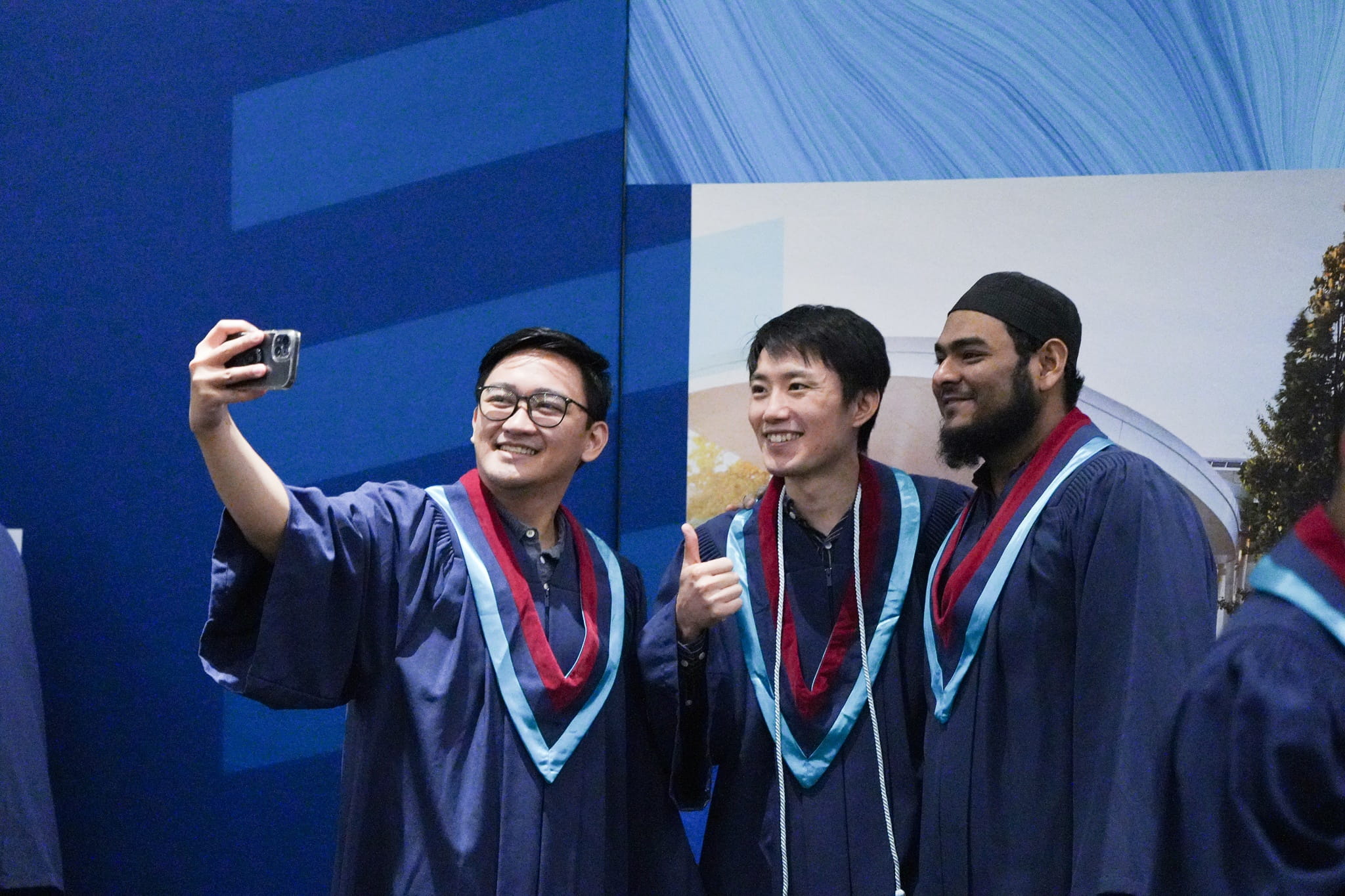 Three male students wearing robes and stoles pose for a selfie in front of a Sheridan backdrop.