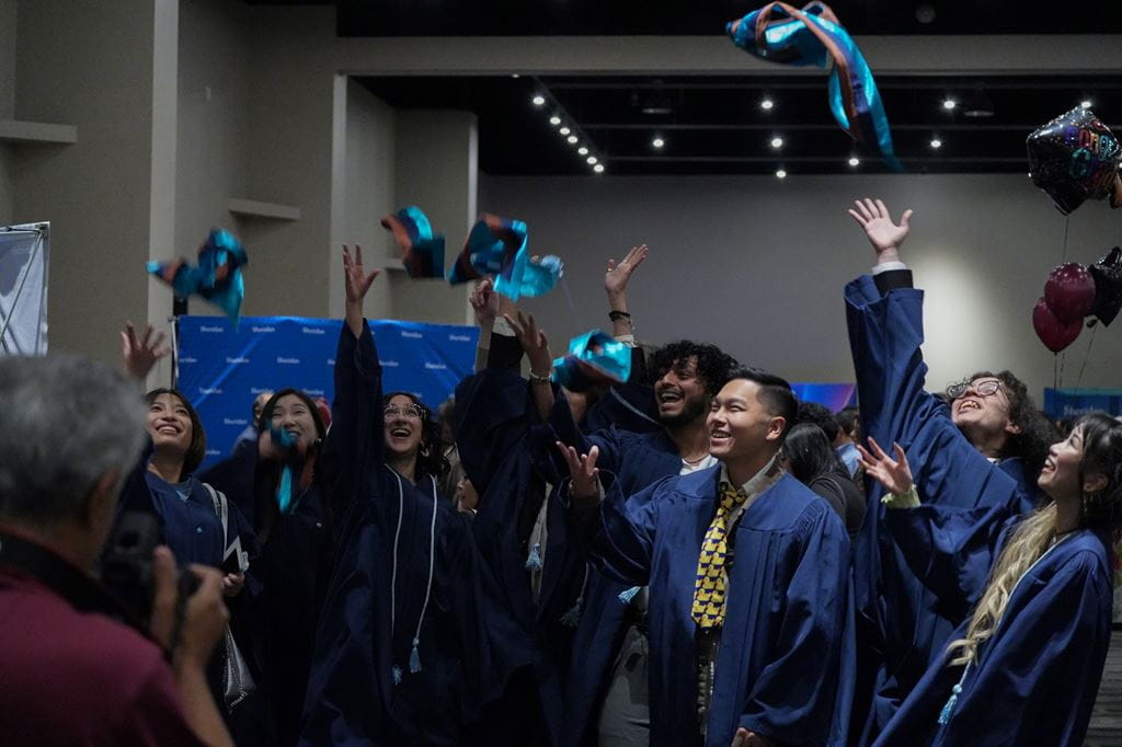 A group of graduating students wearing blue robes toss their stoles into the air.