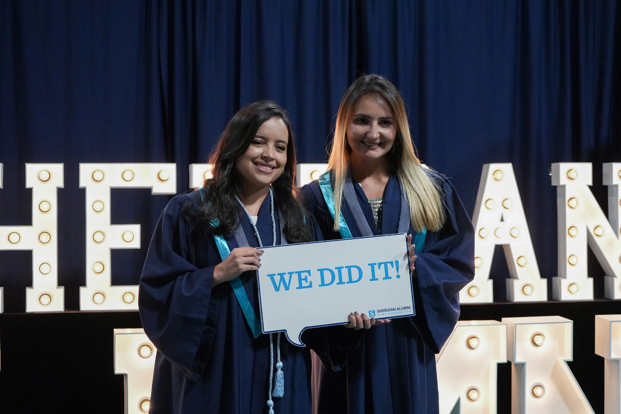 Two female Sheridan students wearing convocation robes hold a sign that says We Did It!