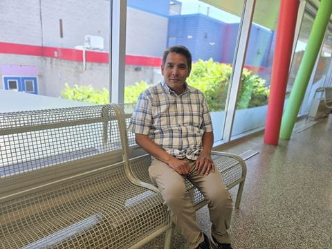 A portrait photo of Daren Costin sitting on a metal bench at Sheridan's Trafalgar Campus with large windows in the background