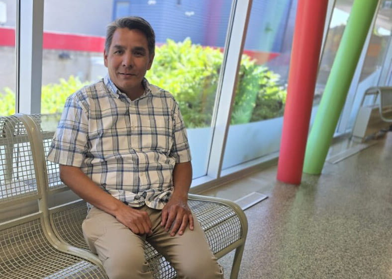 A portrait photo of Daren Costin sitting on a metal bench at Sheridan's Trafalgar Campus with large windows in the background