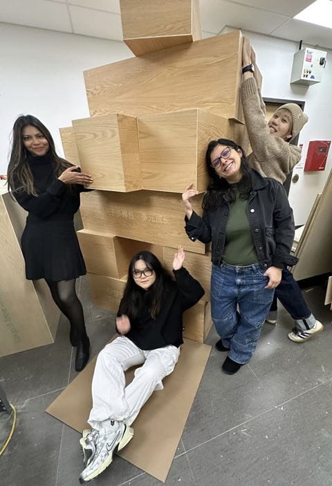 Four Visual Merchandising Design students stand and sit around the large wooden blocks they created for Jenga Roots