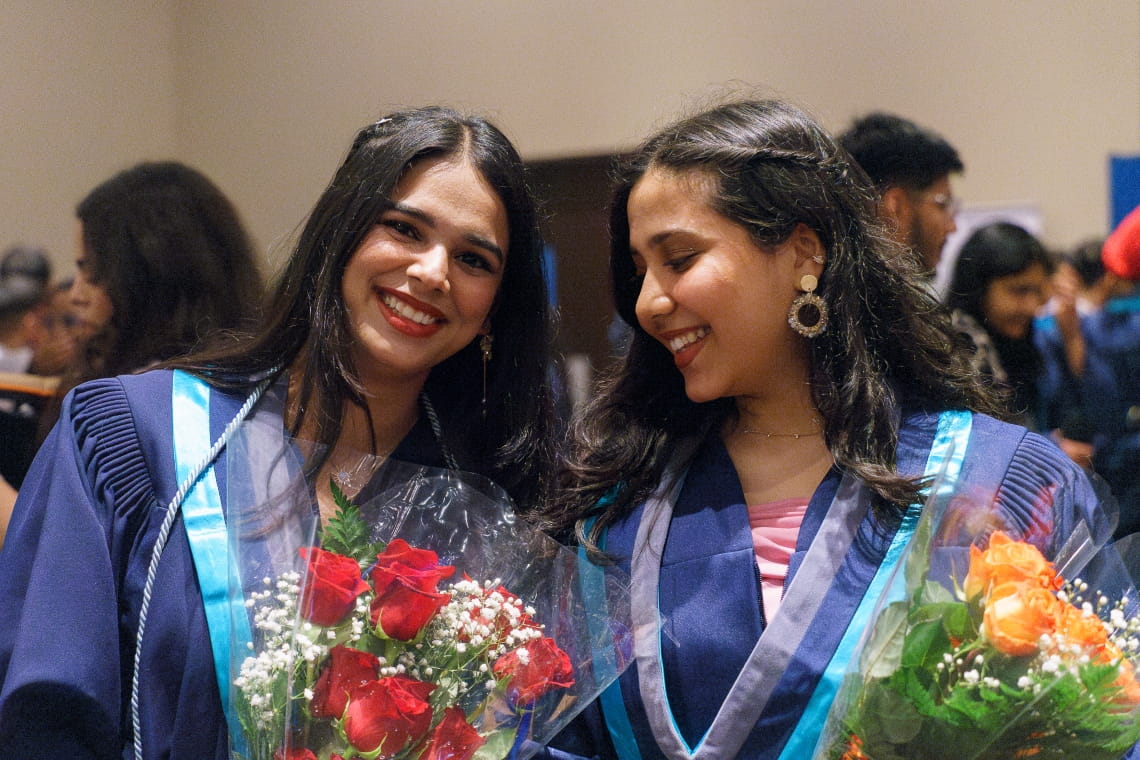 Two women wearing grad gowns and holding flower bouquets stand together and smile.
