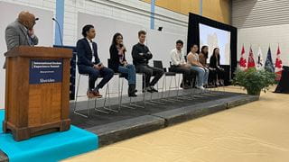 A panel of students seated side-by-side across a stage. The moderator stands to the left behind a podium.