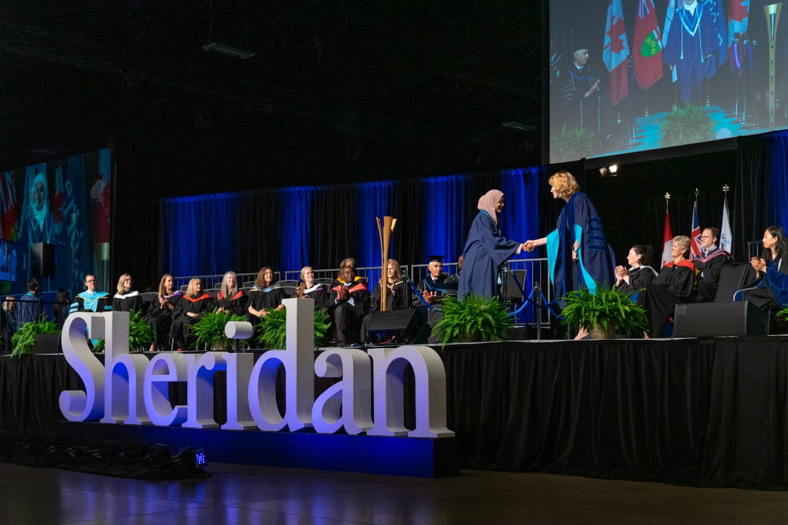 President Janet Morrison shakes the hand of a graduate who is crossing the convocation stage. The platform party sits on chairs in the background.