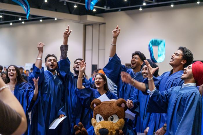 A group of students wearing convocation robes toss their sashes in the air while celebrating. Mascot Bruno the Bear is in the foreground.