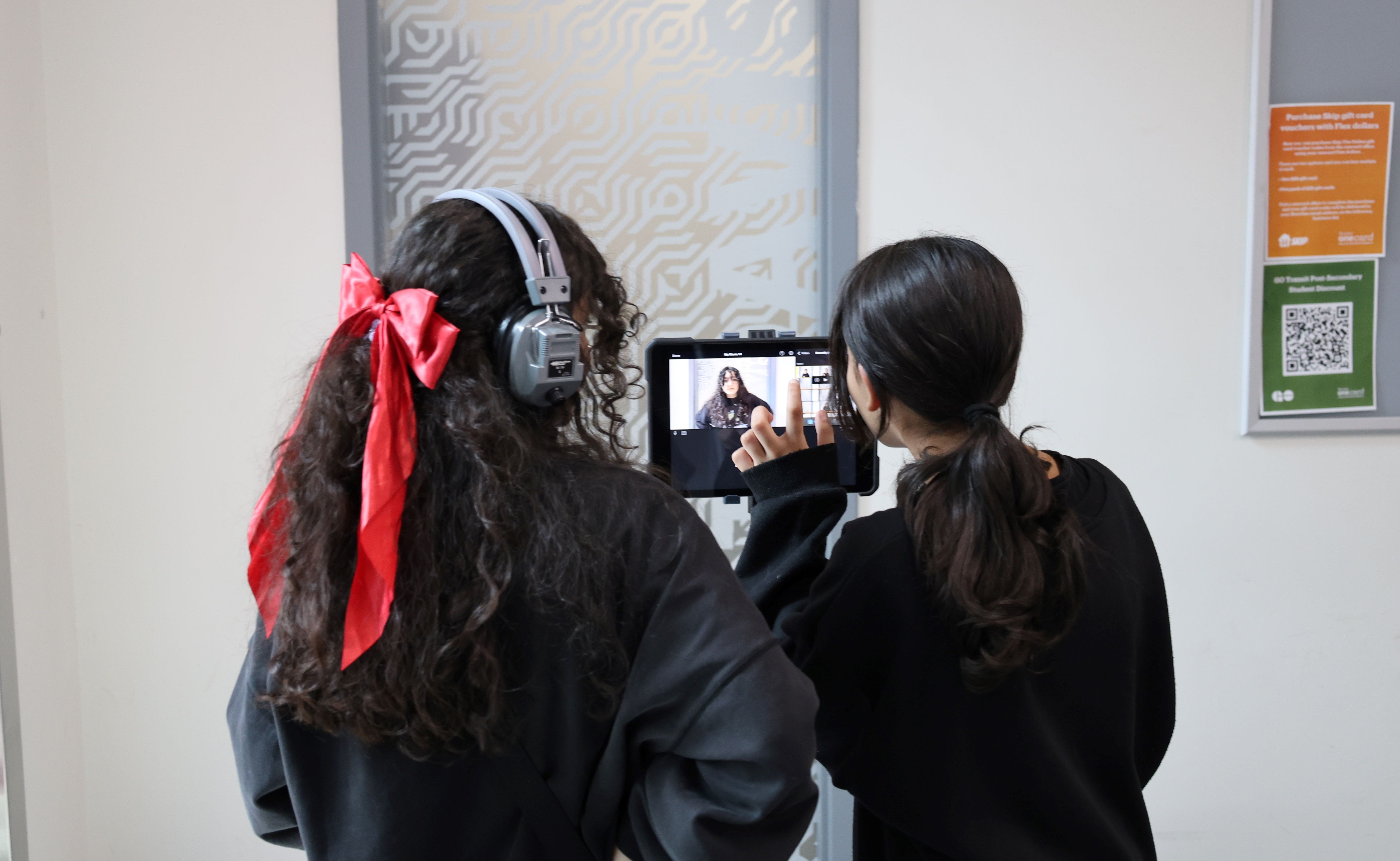 Two students stand with their backs to the camera, facing the screen of a video camera.