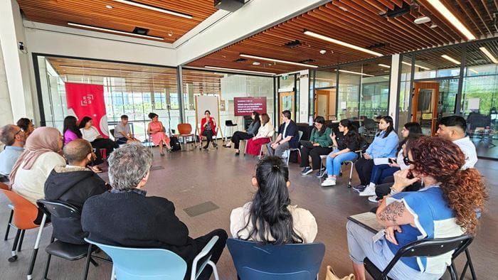 A large group of people sit on chairs in a circle in a room with many large windows.
