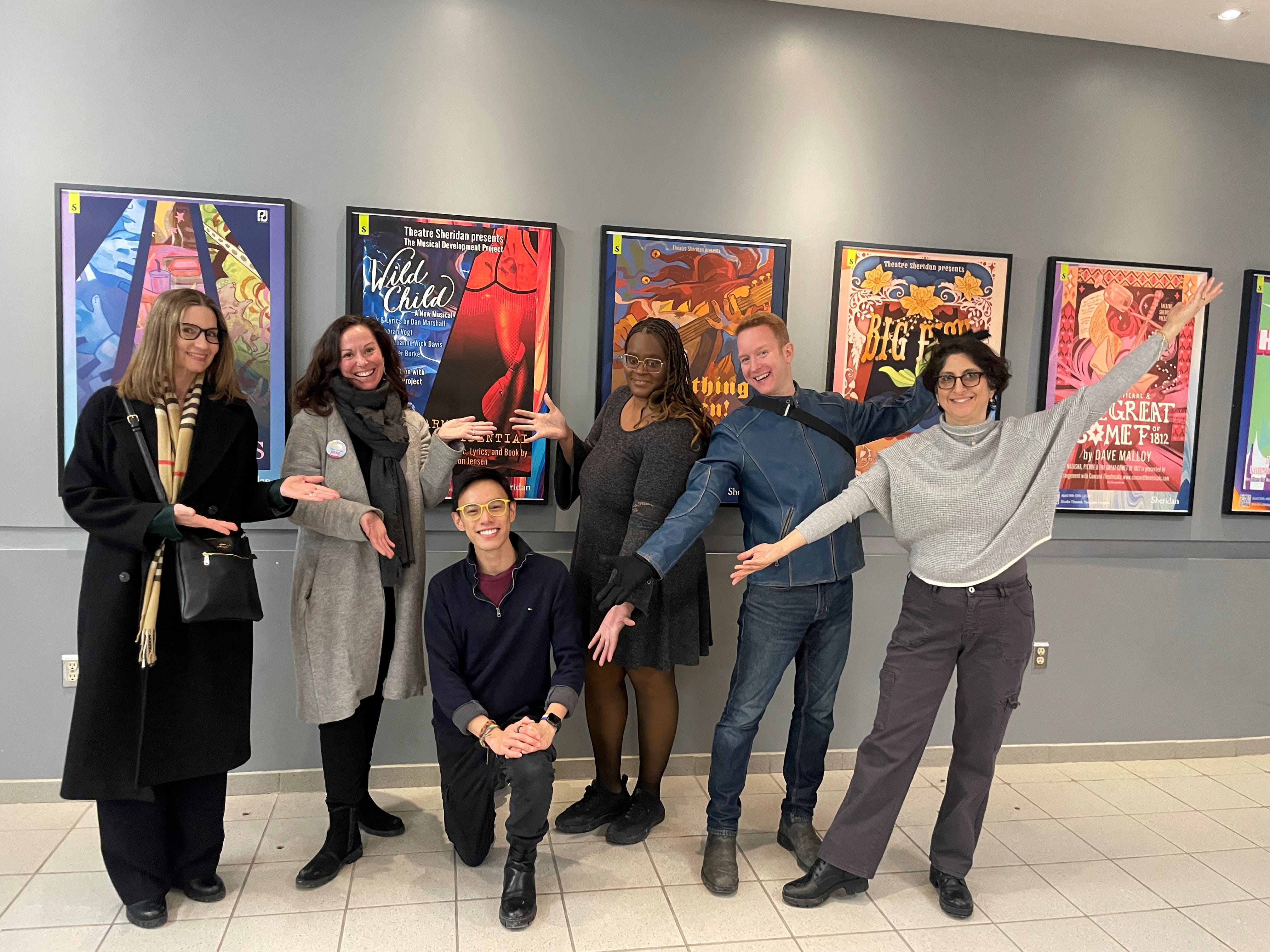 Six people pose for a photo. One person is kneeling and the five people standing are gesturing towards him. They are standing in front of a group of theatre posters on the wall.