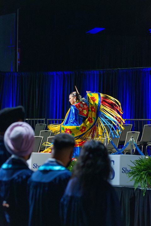 An Indigenous woman in a colourful ribbon dress dances on the convocation stage.