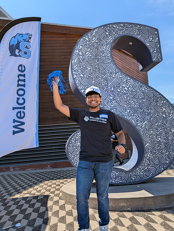 A Sheridan student stand cheerfully in front of a welcome flag and a large Sheridan 'S'