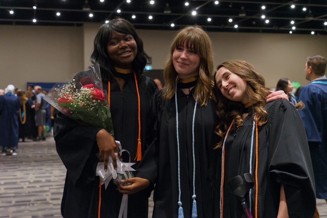 Three students wearing black grad gowns, one is holding flowers, smile for a photo.