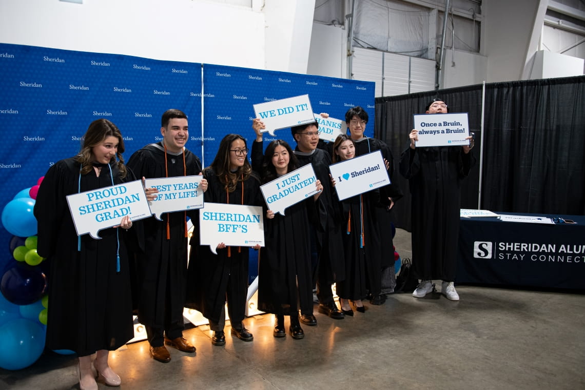 Eight Sheridan grads wearing grad gowns hold signs celebrating their graduation in front of a blue Sheridan banner.