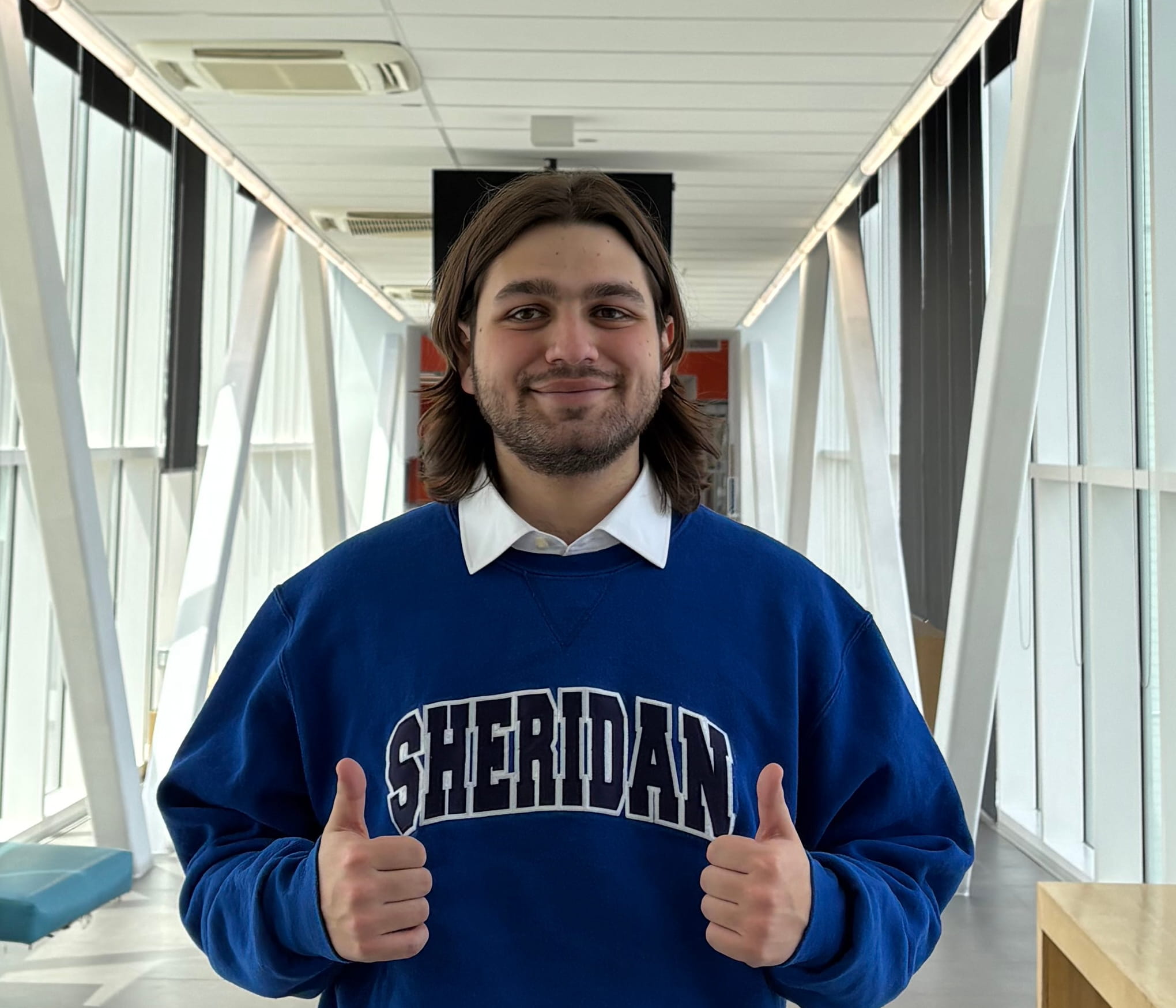 Valedictorian Samuel Heyman stands in a hallway wearing a blue Sheridan hoodie and holding two thumbs up.