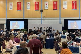 Looking across the crowd at a group of panelists seated side-by-side. Large screens sit at each end of the group.
