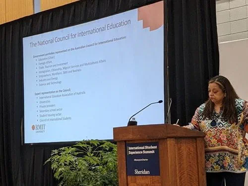 Roopa Desai Trilokekar standing behind a podium that reads 'International Student Experience Summit'. A large projection screen to her left displays her presentation.
