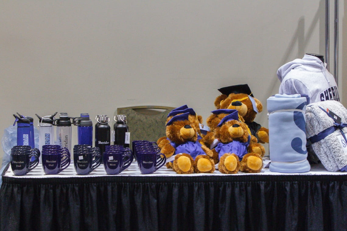 A table displays items with Sheridan swag: mugs, water bottles, teddy bears, blankets and a hoodie.