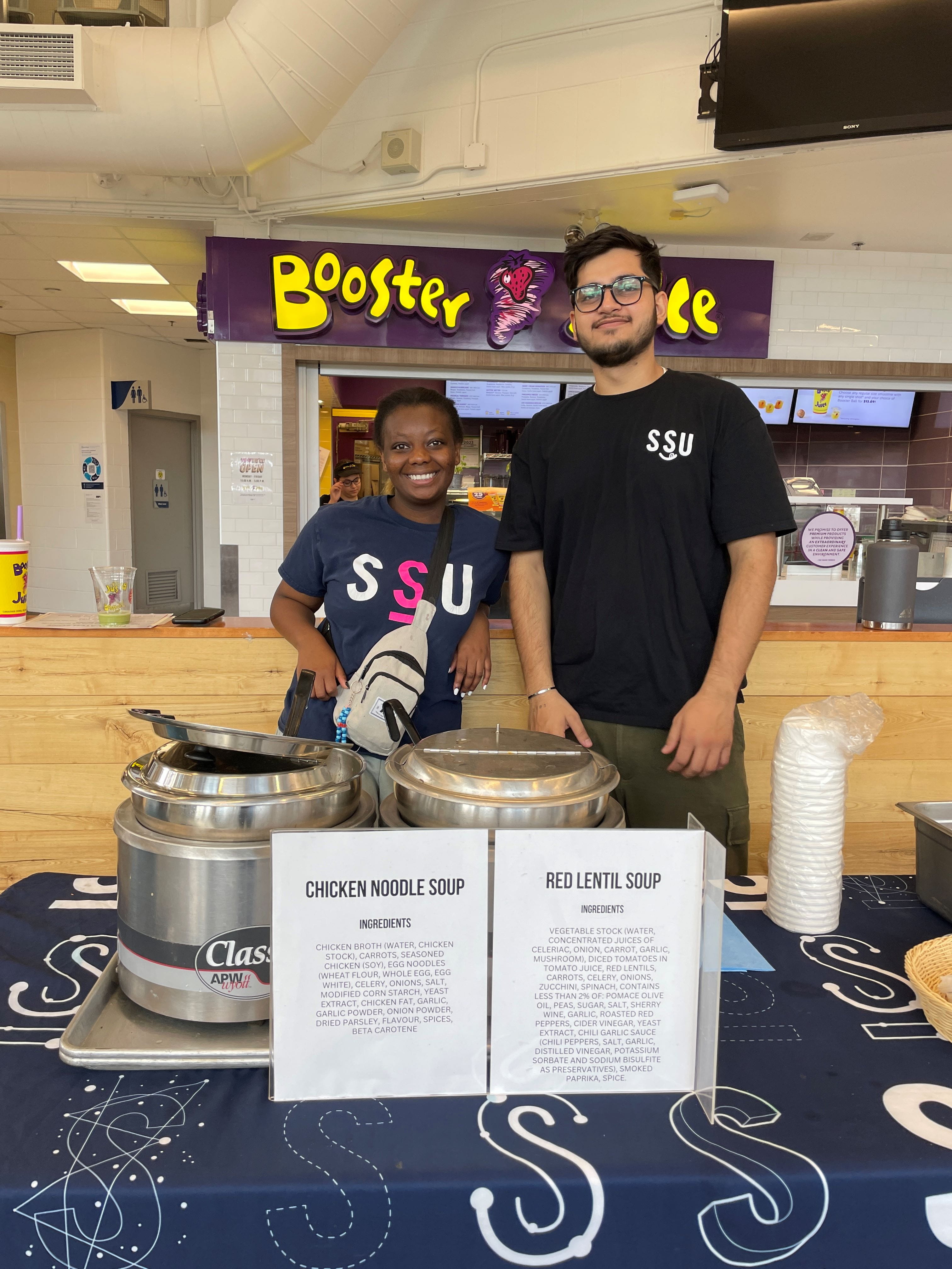 Two students wearing SSU shirts stand at a table with two large pots of soup.