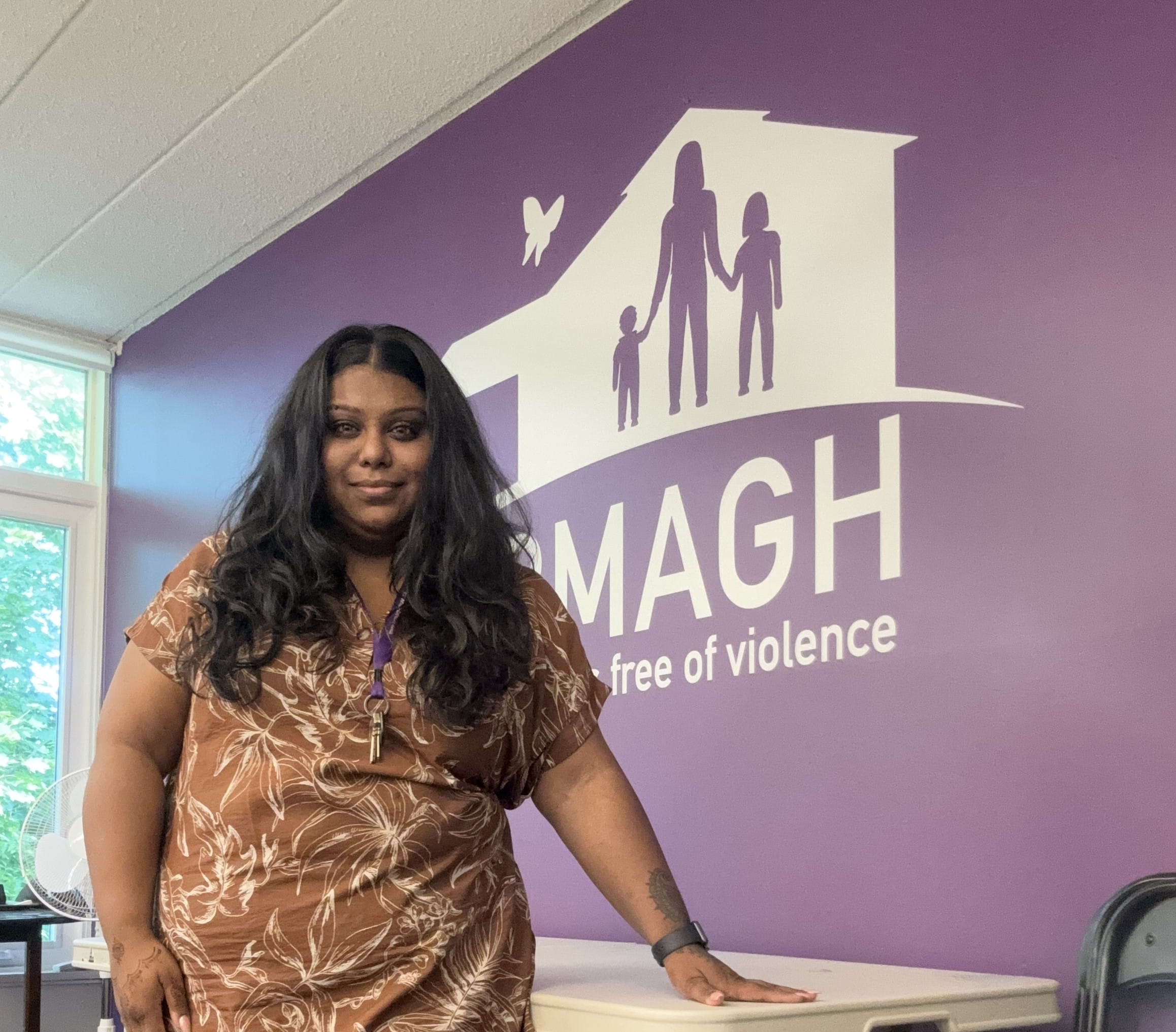 Thanah Sriskantha stands in front of a purple wall depicting the Armagh House logo