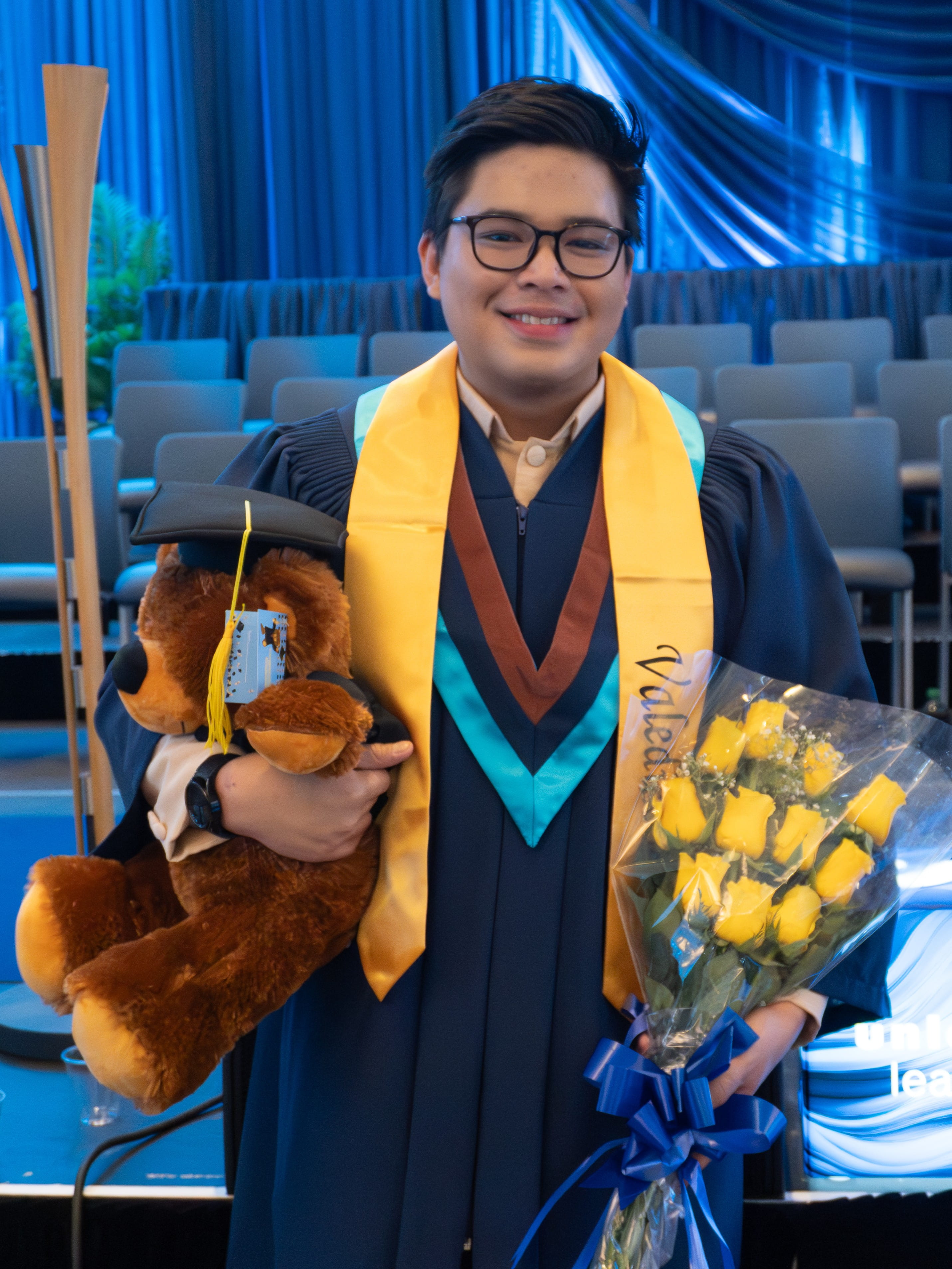 Valedictorian Nathan Josiah Sasing stands in front of a stage holding a teddy bear and yellow roses.
