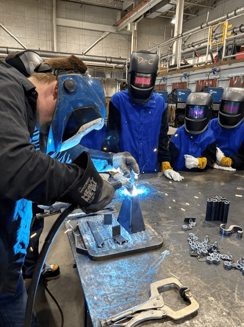 A participant wearing welding safety gear welds metal pieces at a workbench in a welding lab while several other participants in welding helmets observe. Tools, clamps and metal components are arranged on the table, and bright welding sparks are visible.
