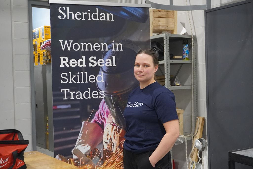Kelsey Annan stands in a Sheridan skilled trades lab with a Women in Red Seal Skilled Trades promotional banner in the background