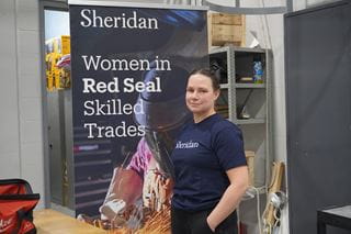Kelsey Annan stands in a Sheridan skilled trades lab with a Women in Red Seal Skilled Trades promotional banner in the background