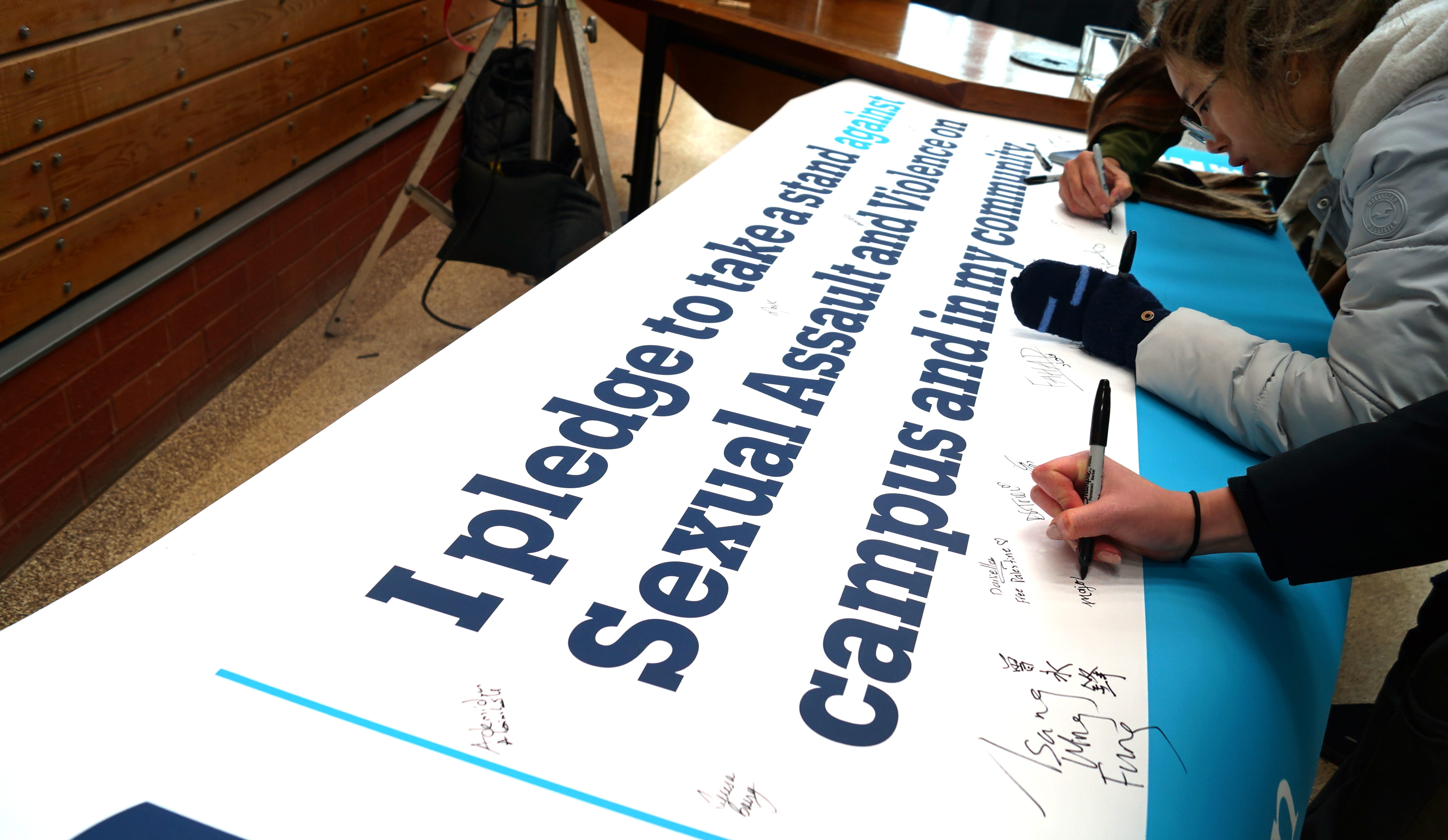 People use markers to sign a banner on a table that says I pledge to take a stand against sexual assault and violence on campus and in my community.