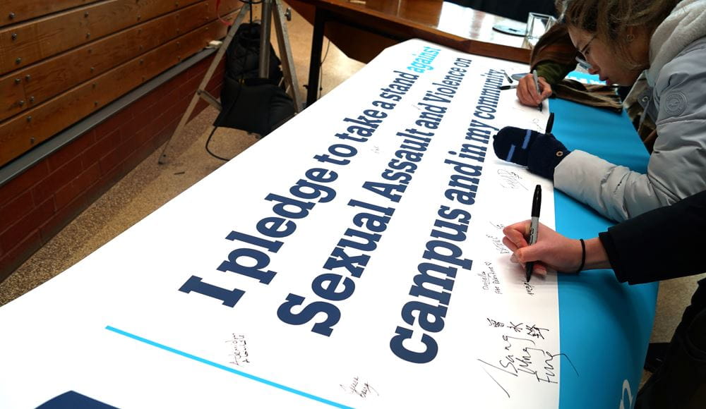 People use markers to sign a banner on a table that says I pledge to take a stand against sexual assault and violence on campus and in my community.