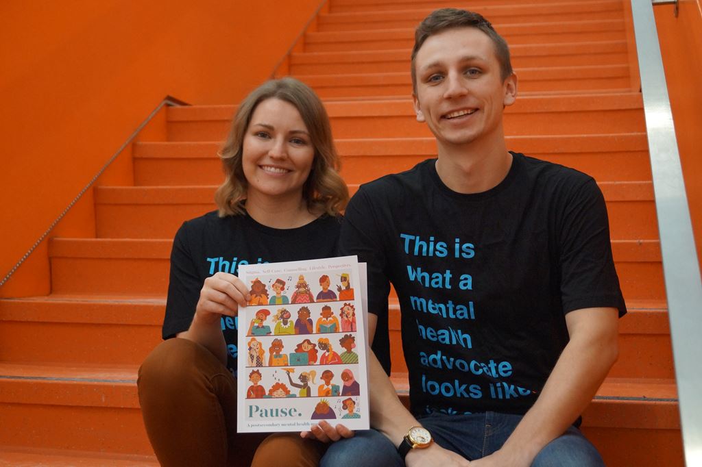 Brittany Tapper and Ben LeBlanc sitting on the orange stairs at Sheridan's Hazel McCallion Campus holding up Pause. Magazine