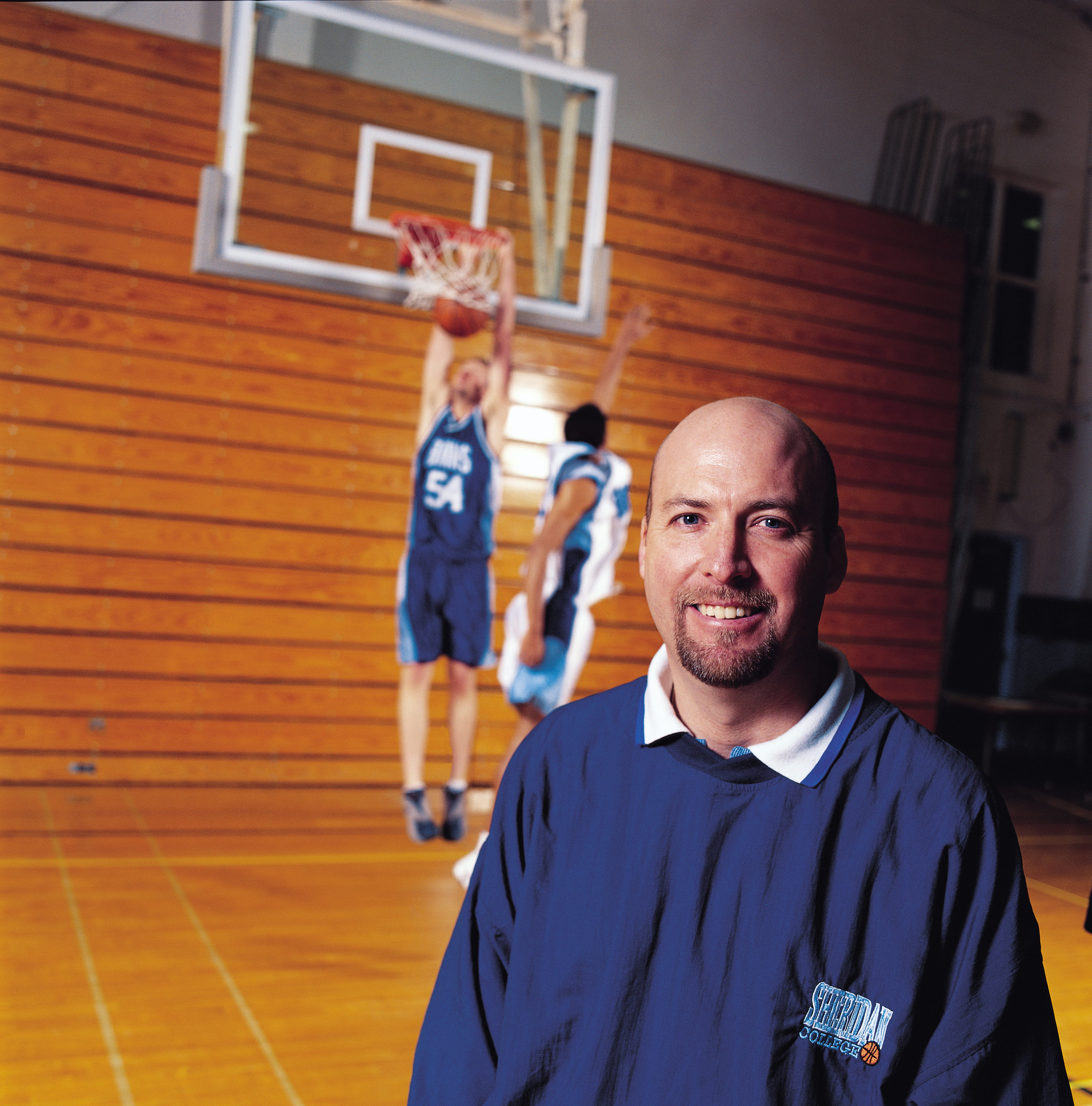 Sheridan athletic director Jim Flack stands in the Trafalgar Road Campus gymnasium while two basketball players play in the background
