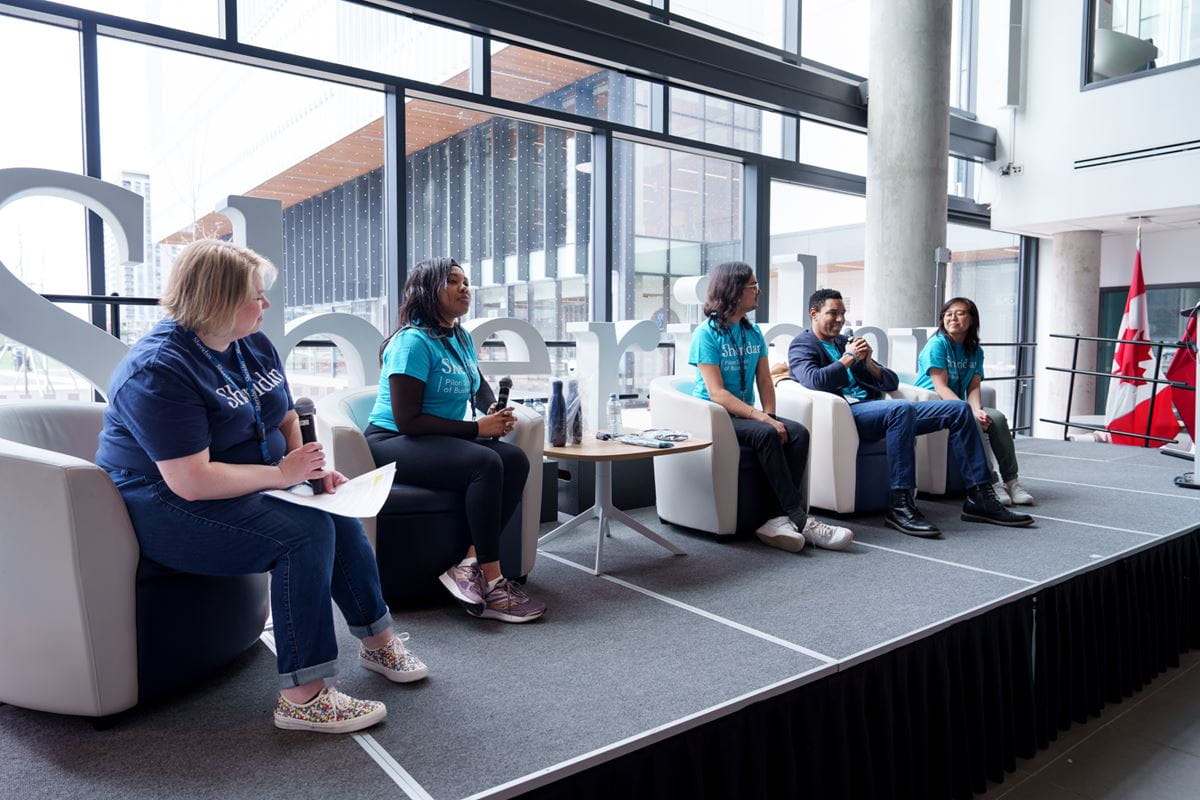 Five people sit on chairs on a stage in front of a window and one speaks into a microphone.