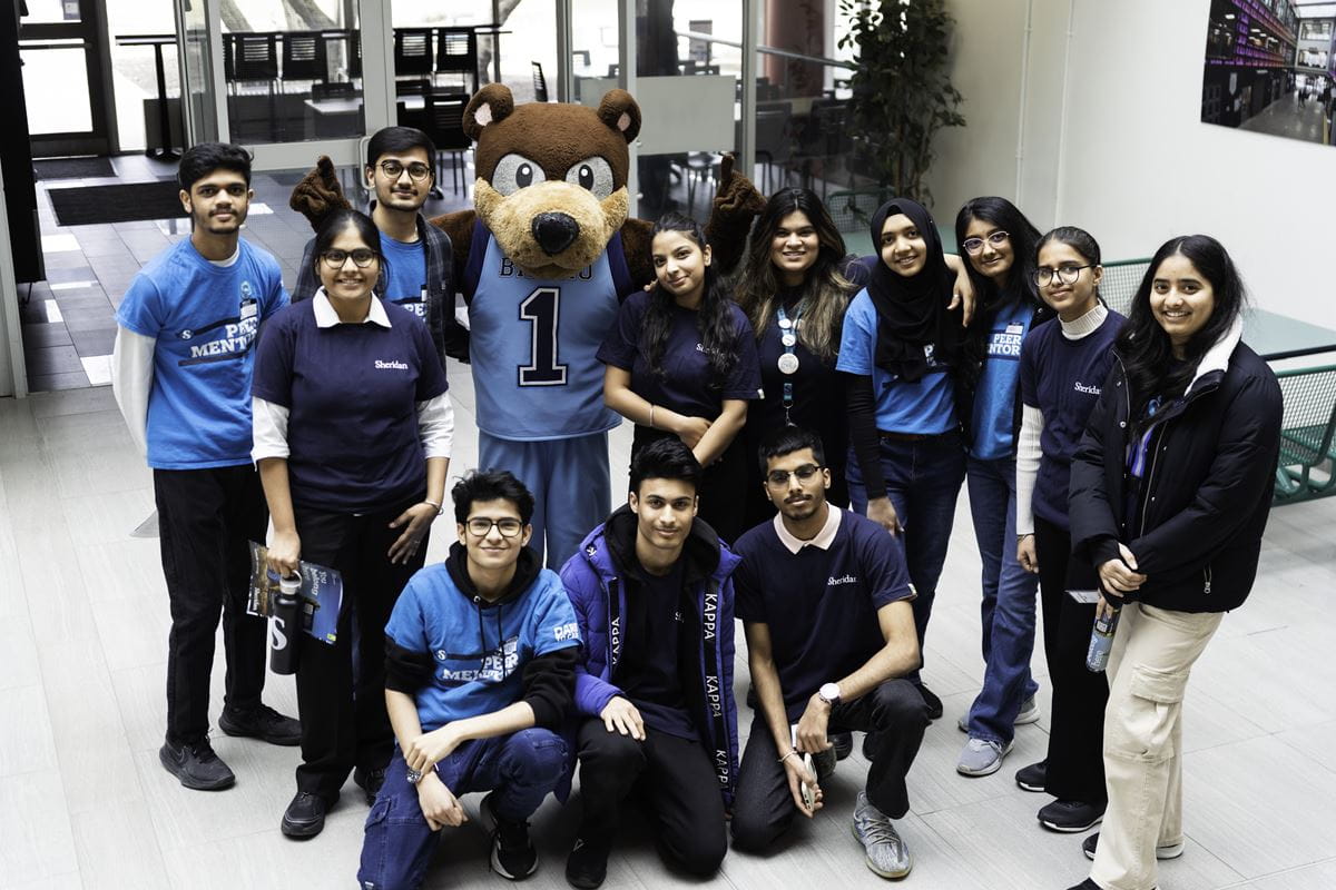 A group of students mostly wearing blue Sheridan branded shirts, gathers around bear mascot Bruno.