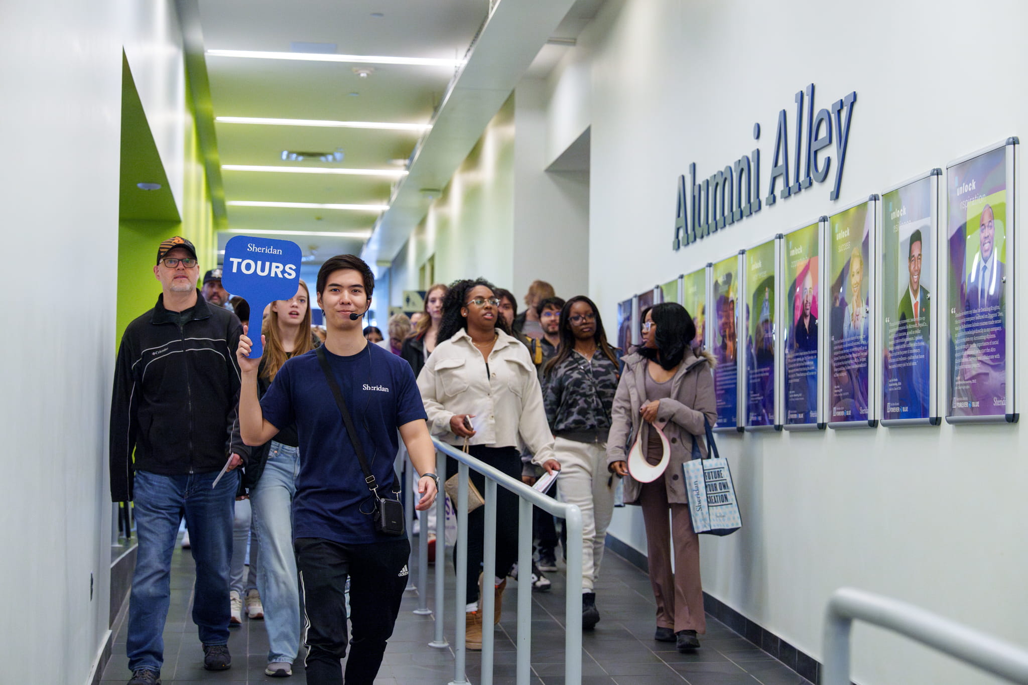 A small group of people walk down a hallway past photos on the wall with a sign that says Alumni Alley.