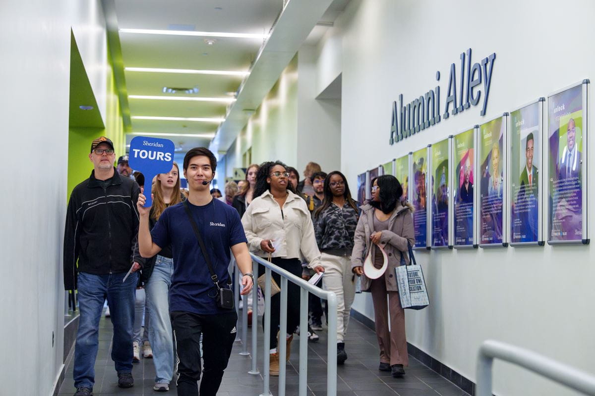A small group of people walk down a hallway past photos on the wall with a sign that says Alumni Alley.
