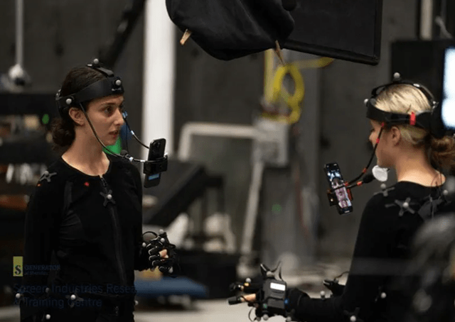Two researchers face each other while wearing black body suits with sensors attached and phones displayed in front of their faces