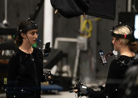 Two researchers face each other while wearing black body suits with sensors attached and phones displayed in front of their faces