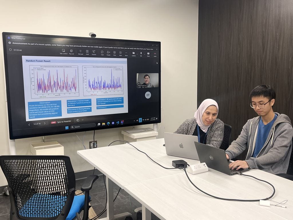 Sheridan professor Haya El Ghalayini sits at a table beside a student and in front of a large screen that displays research data