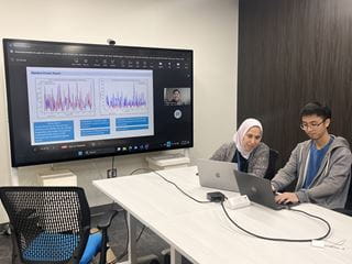 Sheridan professor Haya El Ghalayini sits at a table beside a student and in front of a large screen that displays research data