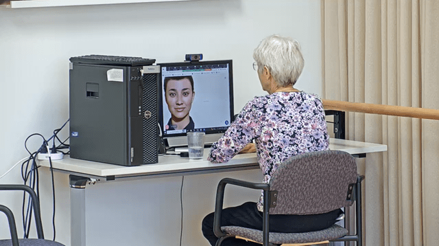An older adult sits in front of a computer and interacts with a virtual human displayed on the monitor