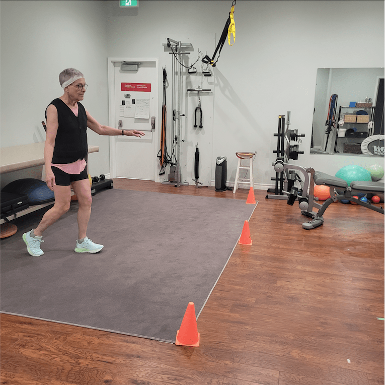 An older adult moves while standing on a mat inside a gym space.