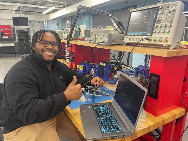 Sheridan electrical engineering student Nathan Morgan smiles and gives a thumbs up while sitting at a table next to electronics and a laptop
