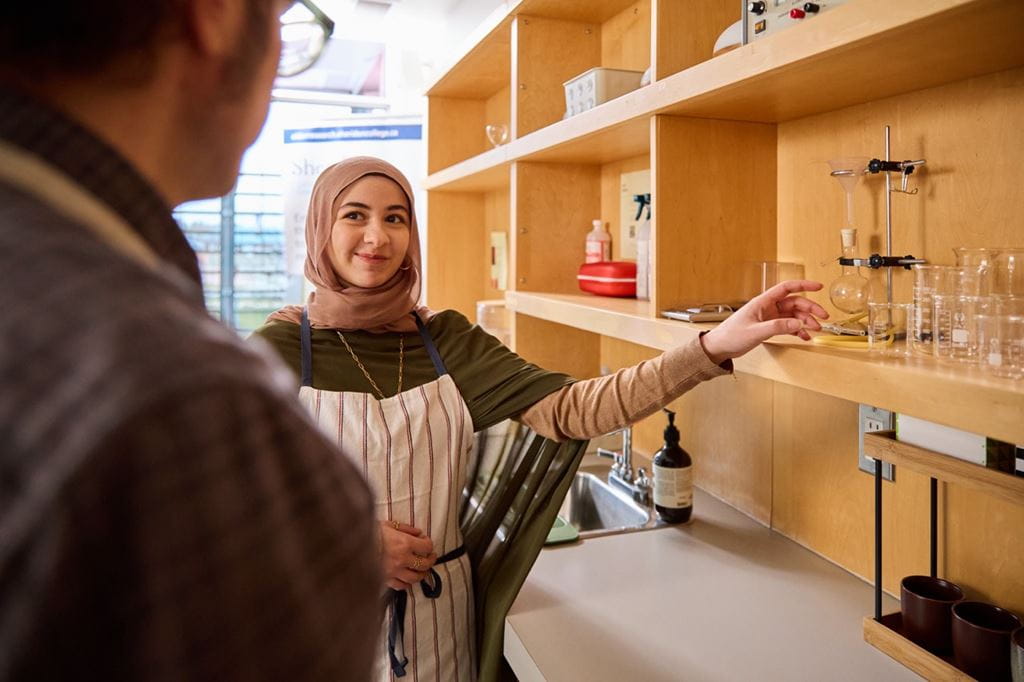 Experiential Design student Samira Elbehairy looks at a person while reaching for a glass item on a wooden shelf