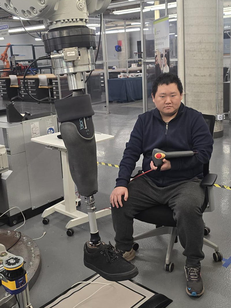 Shengwei Kang sitting next to a prosthetic leg mounted on a robotic arm
