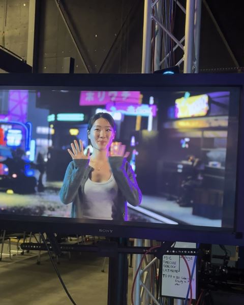 A production monitor on a studio set displays a person standing on a neon-lit urban street scene. The person is centered on the screen with both hands raised near shoulder height, while colourful pink, blue, and yellow signs glow in the blurred background. The monitor is surrounded by metal truss structures, cables, and backstage equipment, revealing the behind-the-scenes filming environment